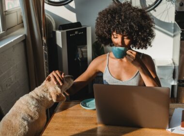 Black woman freelancer drinking coffee by her computer with dog