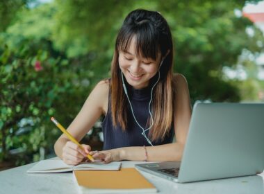 Happy Asian woman student writing on computer with headphones in ears